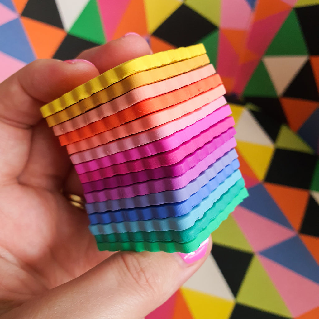 Hand holding a stack of colourful rainbow polymer clay swatches. Background is brightly coloured and geometric.